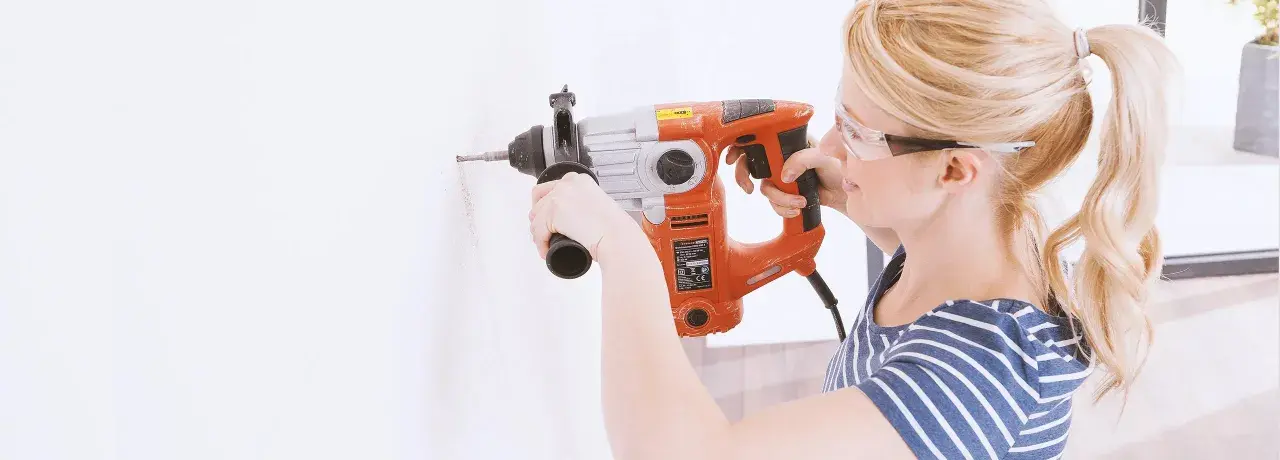 Frau bohrt mit Schlagbohrmaschine ein Loch in die Wand. Sie trägt Schutzbrille.