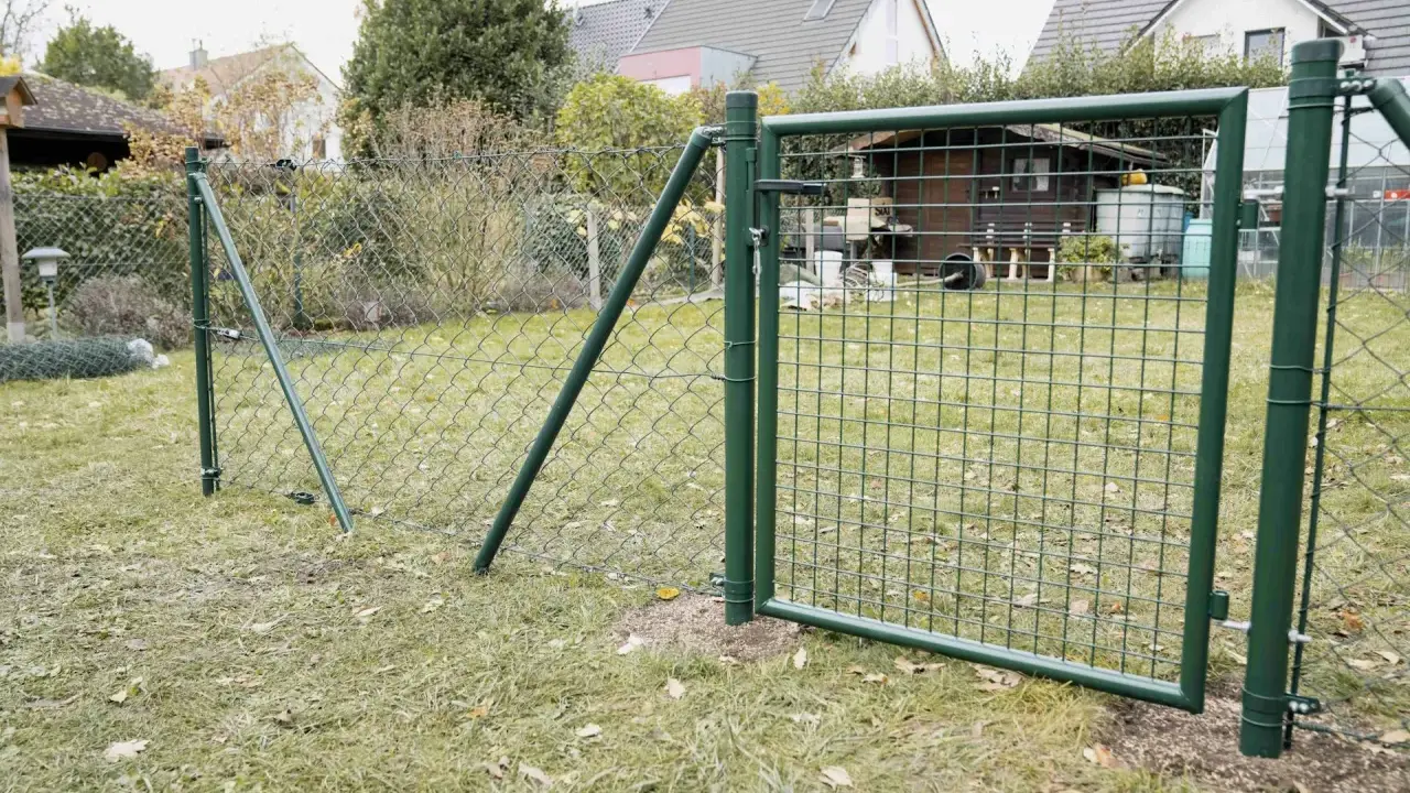 Ein grüner Maschendrahtzaun wird gespannt, mit einem Tor, das offen steht. Im Hintergrund sind Häuser und ein Schuppen zu sehen.