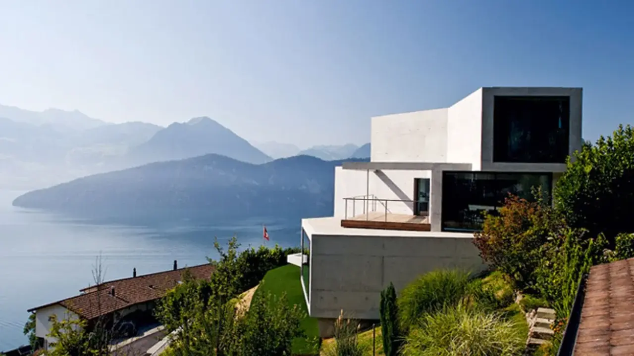 Modernes Haus in Hanglage mit Blick auf einen See und Berge. Ein traditionelles Haus liegt tiefer am Hang.