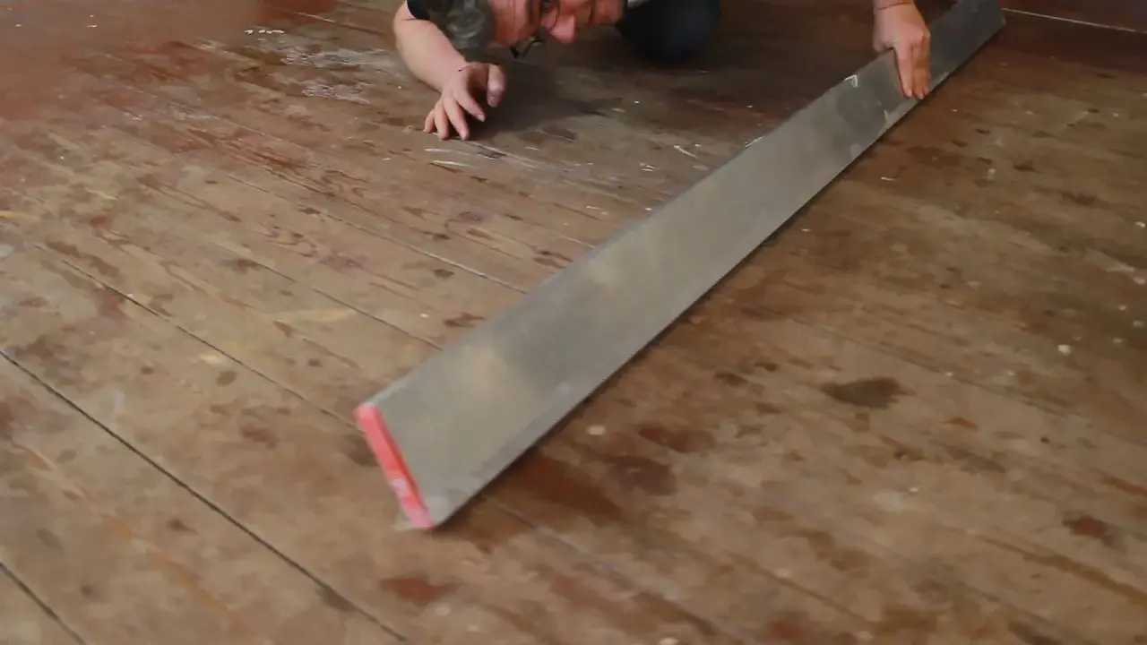 Mann verlegt Dielenboden, hält ein langes Metallwerkzeug, um die Bretter auszurichten.