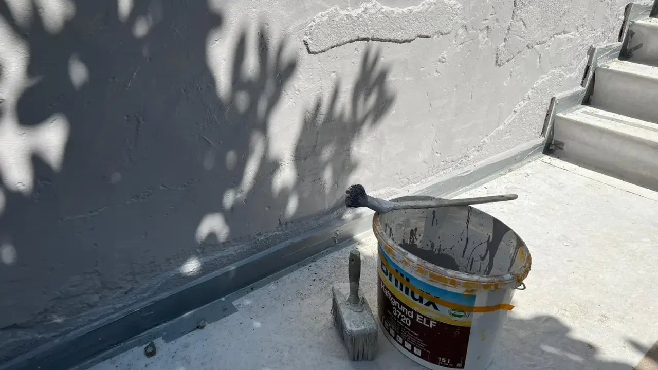 Werkzeug und Farbeimer bereit zum Ausbessern des Sockelputzes. Schatten von Blättern fallen auf die Wand.