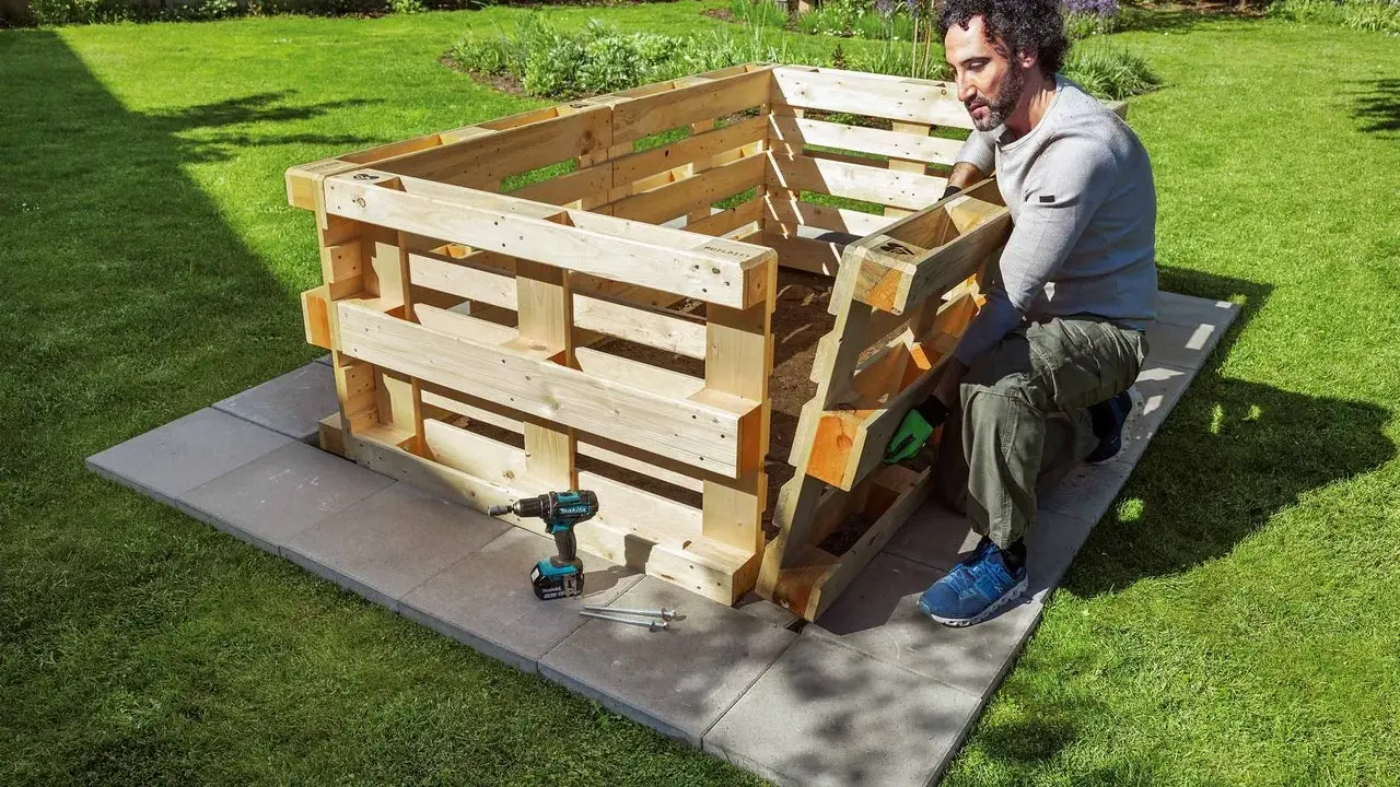 Mann baut ein **Hochbeet aus Paletten** im Garten. Werkzeug und Material liegen bereit.