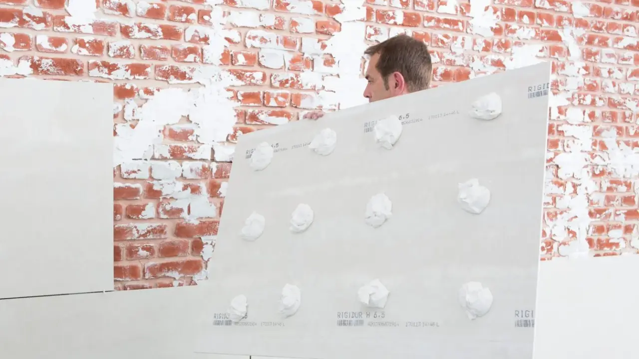 Mann hält Gipskartonplatten mit Klebepunkten, um sie auf unebener Wand zu befestigen.