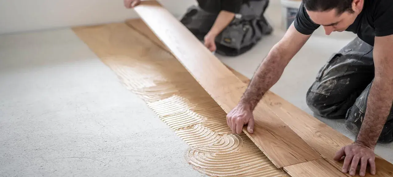 Handwerker verlegen sorgfältig Parkett. Kleber wird auf den Boden aufgetragen, bevor die Holzplanken präzise platziert werden.