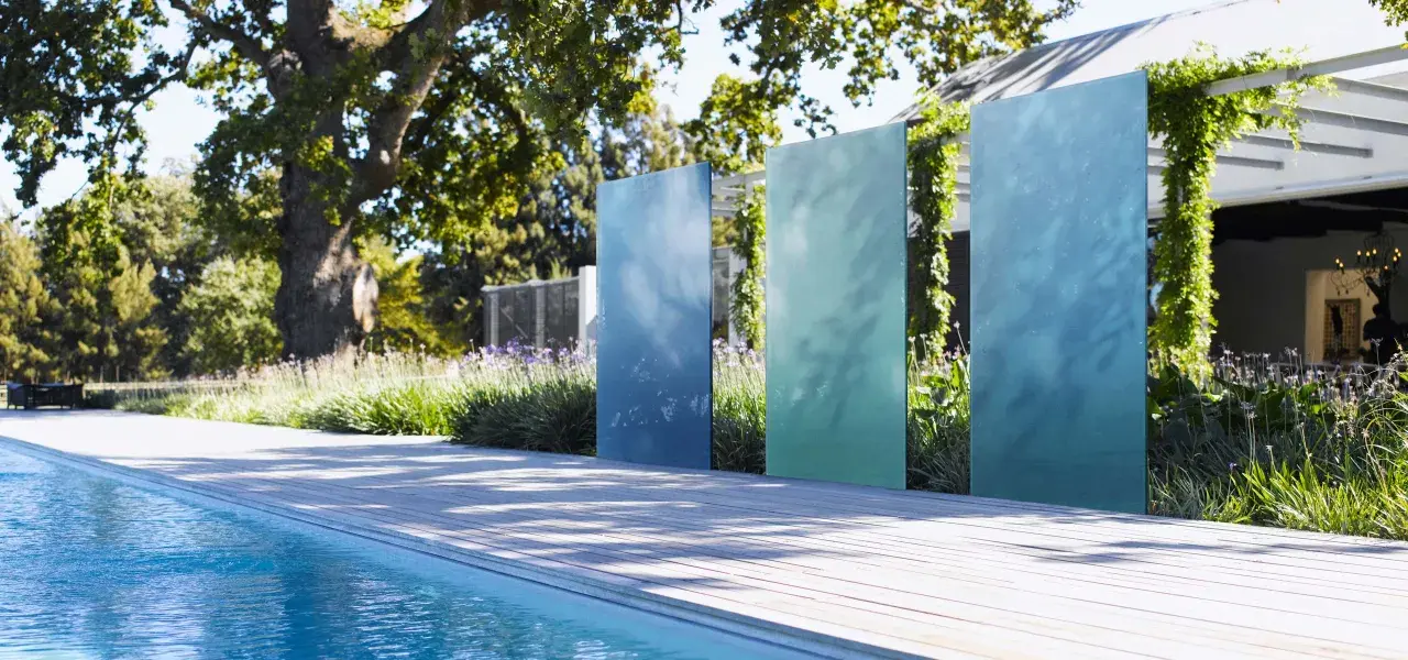 Ausgefallener Sichtschutz Garten selber machen: Vertikale, blaue und grüne Metallplatten vor üppiger Bepflanzung und Pool.