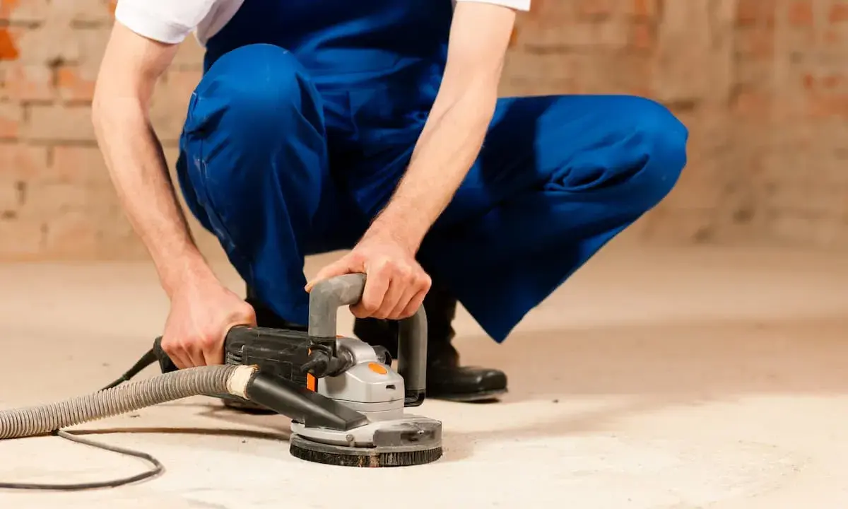 Mann in blauer Latzhose schleift mit einer Maschine den Estrich. Staub wird abgesaugt.