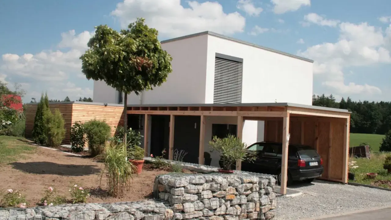 Modernes, weißes Haus mit Holzcarport und Gabionenmauer. Ein Beispiel, wie man ein low budget haus bauen kann, das dennoch stilvoll aussieht.