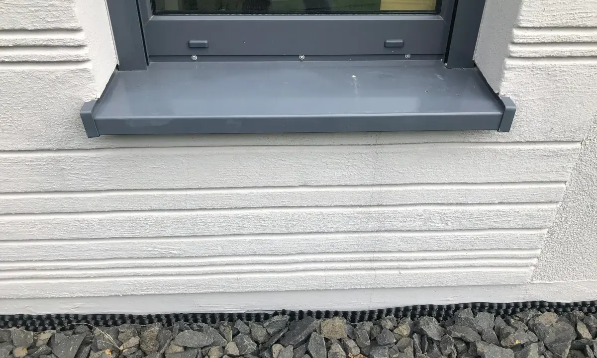Graue Fensterbank mit leichter Neigung, die Regenwasser ableitet. Darunter weiße Holzverkleidung und grauer Schotter.