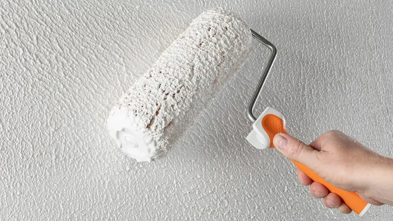 Eine Hand mit orangefarbenem Griff hält eine Farbrolle, die weißen Putz auf eine strukturierte Wand aufträgt. Der Anschluss Putz an Holz wird hier perfektioniert.