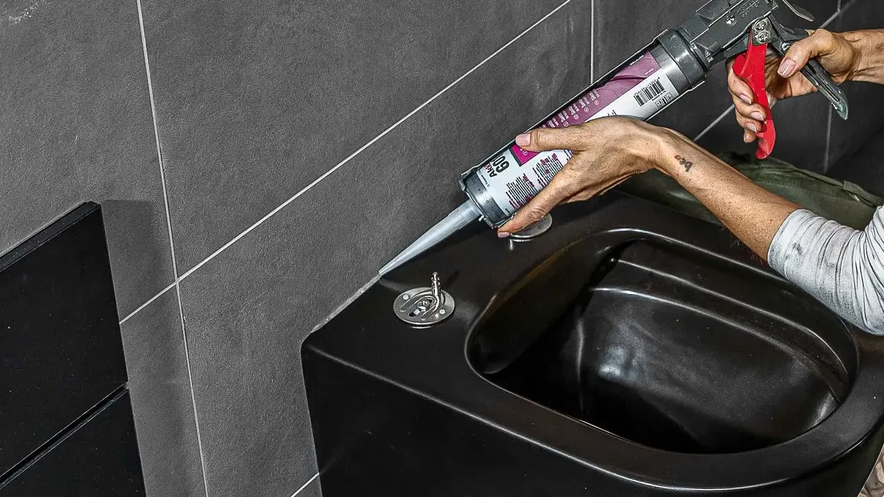 Person erneuert silikonfugen am schwarzen Bidet mit einer Kartuschenpistole.
