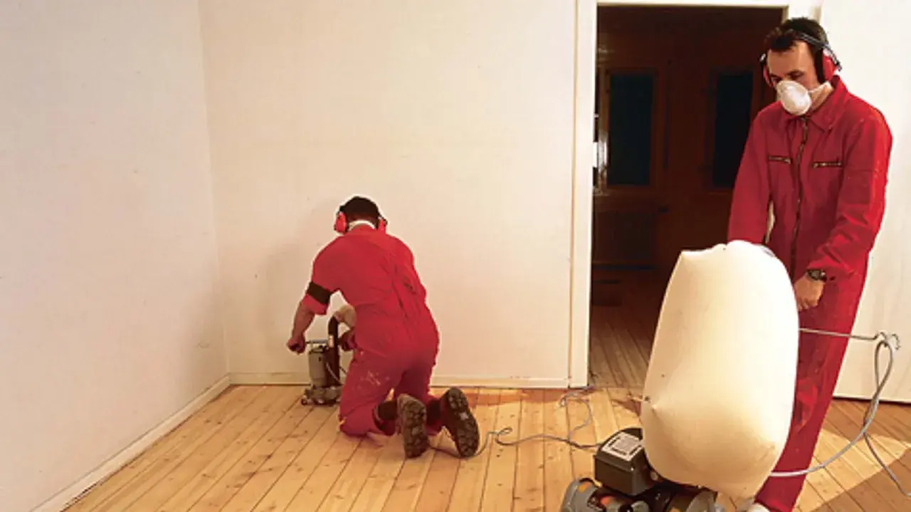 Zwei Männer in roten Overalls schleifen Dielen ab. Einer kniet und bedient eine Schleifmaschine, der andere steht und hält einen Staubfangsack.