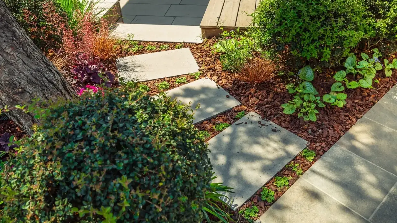 Schritt für Schritt: Gehwegplatten verlegen in einem üppigen Garten mit Rindenmulch und verschiedenen Pflanzen.