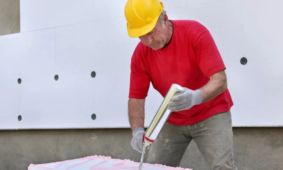 Mann mit gelbem Helm trägt pinken Kleber auf Styropor auf. Er bereitet sich vor, Styropor mit Fliesenkleber zu kleben.