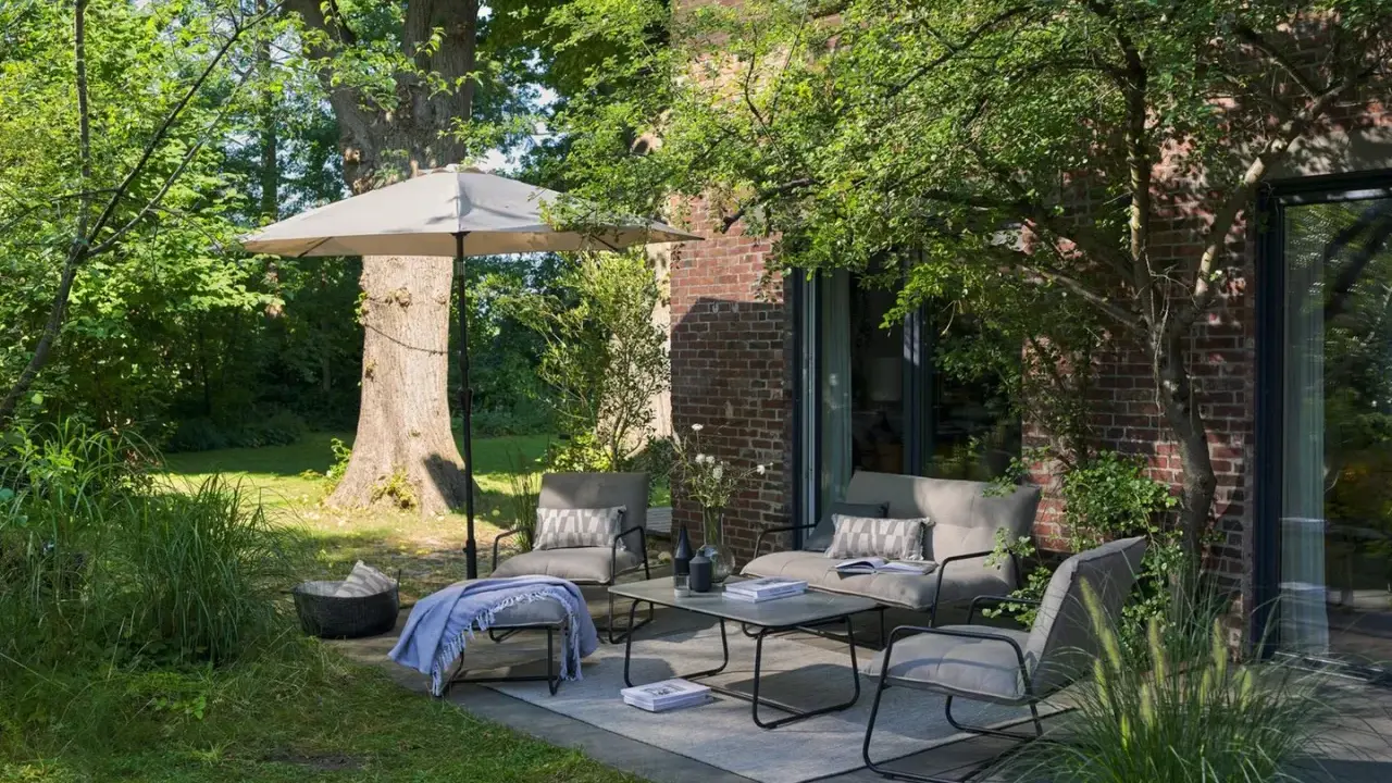 Gemütliche kleine Terrasse gestalten mit bequemen Sitzmöbeln, Tisch, Sonnenschirm und Kissen. Einladend und grün.