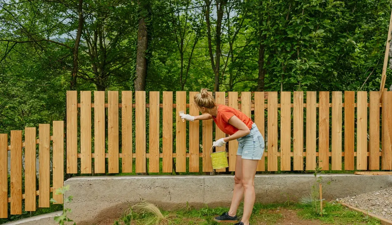 Frau streicht neuen Holzzaun, um ihn vor Witterungseinflüssen zu schützen. Sie setzt den Zaun mit Sorgfalt und Hingabe.