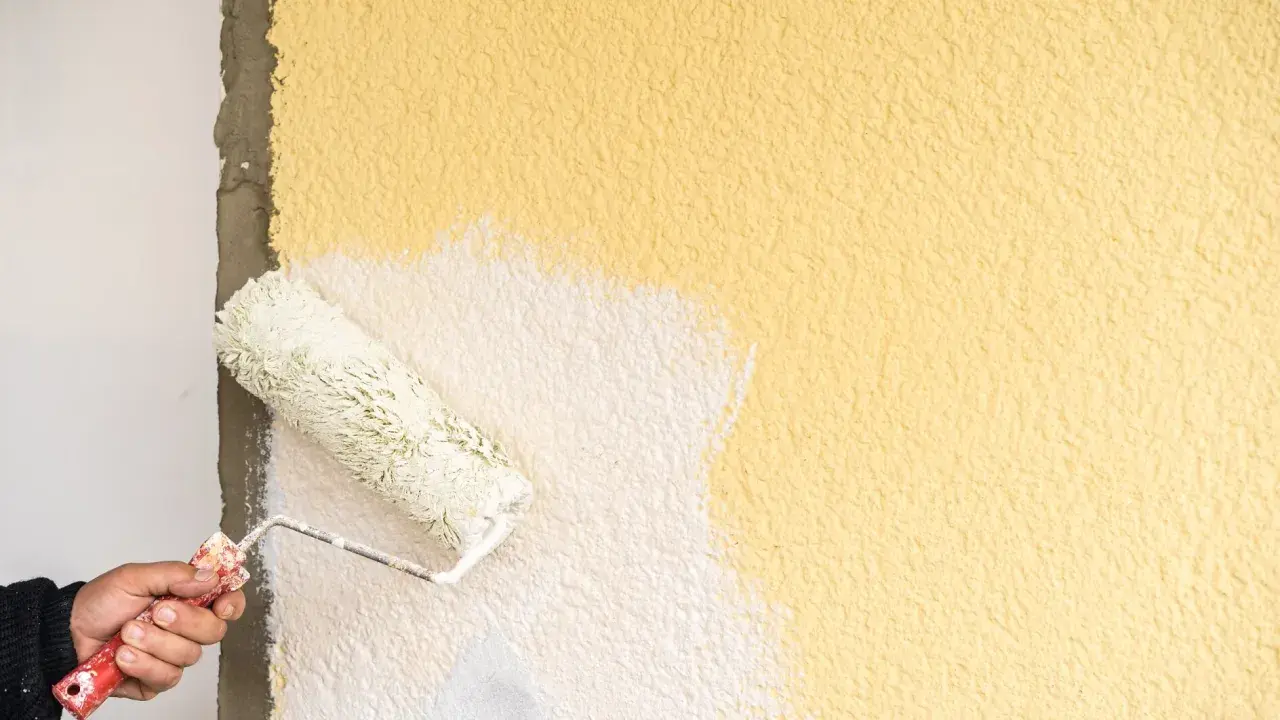 Hand streicht mit einer Farbrolle über eine raue, gelbe Wand. Der Putz wird gerade weiß gestrichen.