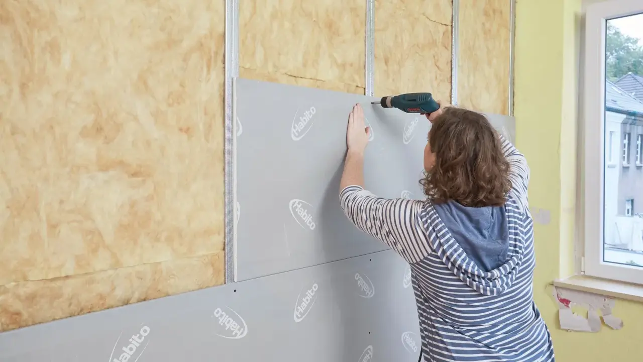 Frau verkleidet Wand mit Rigipsplatten und Schrauber. Isolierung ist sichtbar.