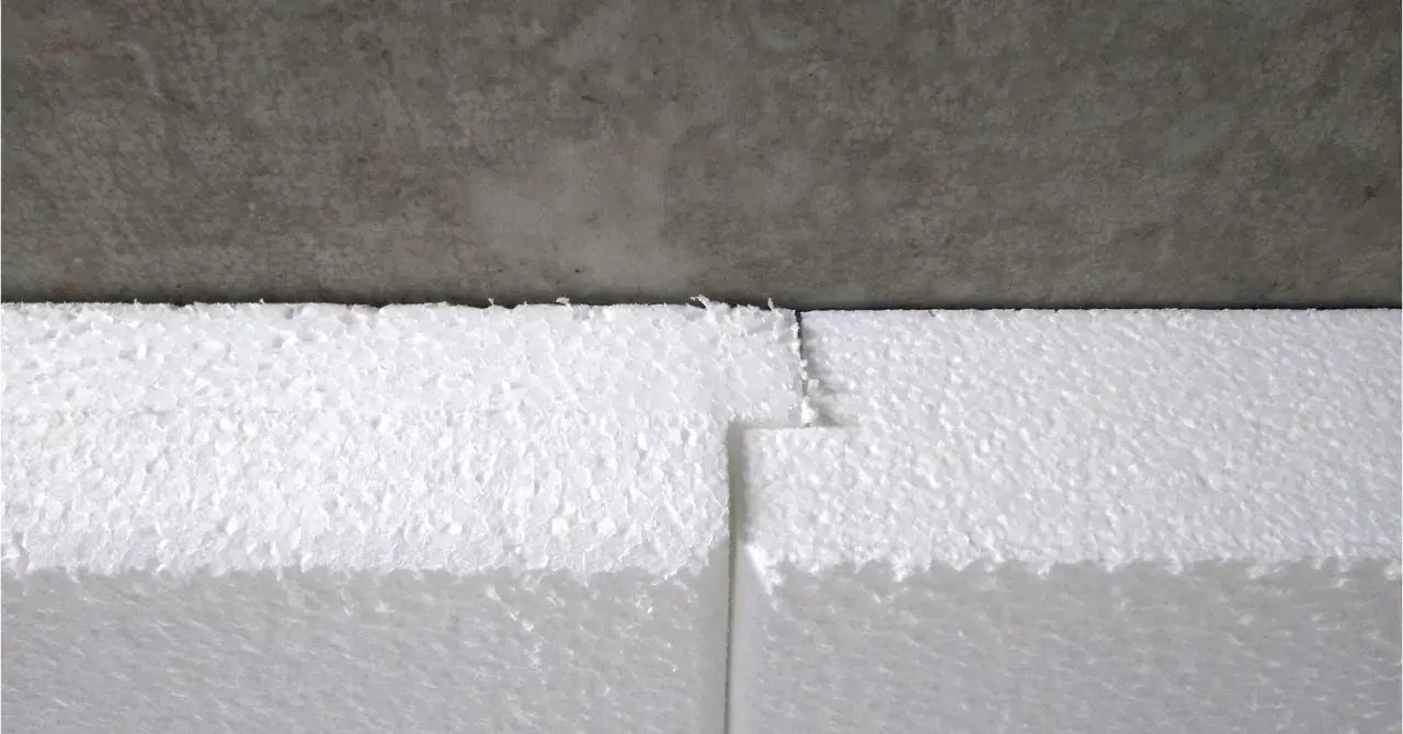Styroporplatten werden zugeschnitten, um ein Kellerfenster zu isolieren. Die weißen Platten sind aneinandergefügt und grenzen an eine graue Betonfläche.