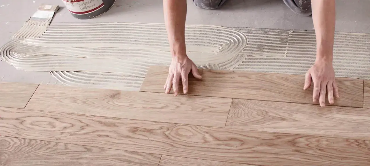 Hände legen Holzplanken auf Kleber, um den Boden zu verlegen.