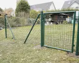 Ein grüner Maschendrahtzaun wird gespannt, mit einem Tor, das offen steht. Im Hintergrund sind Häuser und ein Schuppen zu sehen.
