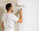 Mann trägt mit einer Malerrolle Rollputz auf eine Wand auf. Die Wand ist halb fertig, die linke Seite ist noch rau, die rechte Seite ist glatt und weiß.
