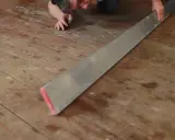 Mann verlegt Dielenboden, hält ein langes Metallwerkzeug, um die Bretter auszurichten.