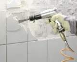 Mit einem Pressluftmeißel werden weiße Fliesen entfernt. Die Wand darunter ist bereits freigelegt und zeigt Mörtelreste.
