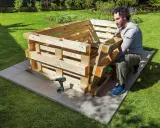 Mann baut ein **Hochbeet aus Paletten** im Garten. Werkzeug und Material liegen bereit.