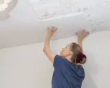 Frau befestigt Styropor-Deckenplatten an einer Decke, um alte, beschädigte Stellen zu verdecken.