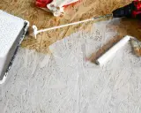 Beim Streichen von OSB-Platten mit weißer Farbe: Ein Farbroller und eine Farbwanne liegen bereit. Handschuhe und ein Akkuschrauber sind ebenfalls zu sehen.