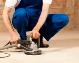 Mann in blauer Latzhose schleift mit einer Maschine den Estrich. Staub wird abgesaugt.