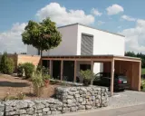 Modernes, weißes Haus mit Holzcarport und Gabionenmauer. Ein Beispiel, wie man ein low budget haus bauen kann, das dennoch stilvoll aussieht.