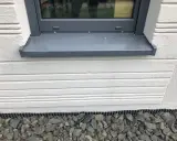 Graue Fensterbank mit leichter Neigung, die Regenwasser ableitet. Darunter weiße Holzverkleidung und grauer Schotter.