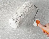 Eine Hand mit orangefarbenem Griff hält eine Farbrolle, die weißen Putz auf eine strukturierte Wand aufträgt. Der Anschluss Putz an Holz wird hier perfektioniert.