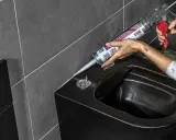 Person erneuert silikonfugen am schwarzen Bidet mit einer Kartuschenpistole.