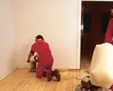 Zwei Männer in roten Overalls schleifen Dielen ab. Einer kniet und bedient eine Schleifmaschine, der andere steht und hält einen Staubfangsack.
