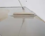Eine Person verteilt mit einem Rakel eine graue Ausgleichsmasse auf einem gefliesten Boden.