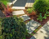 Schritt für Schritt: Gehwegplatten verlegen in einem üppigen Garten mit Rindenmulch und verschiedenen Pflanzen.