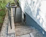 Betontreppe sanieren: Alte Steintreppe mit Geländer führt zu einer Kellertür. Grüne Pflanzen und Kies säumen den Weg.
