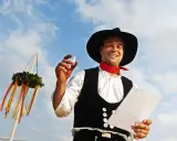 Ein Mann im Zimmermannsgewand hält ein Glas und eine Urkunde. Ein Richtkranz hängt am Himmel. Das ist ein Richtfest!