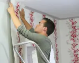 Mann tapeziert Vliestapete mit rotem Blumenmuster. Er steht auf einer Leiter und richtet die Tapete aus, um vliestapete tapezieren fehler zu vermeiden.