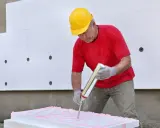 Mann mit gelbem Helm trägt pinken Kleber auf Styropor auf. Er bereitet sich vor, Styropor mit Fliesenkleber zu kleben.
