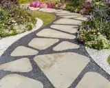 Ein geschwungener Weg aus unregelmäßigen Trittsteinen im Garten, umgeben von bunten Blumen und Kies. Perfekt, um Trittsteine Garten selber machen.