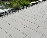 Fertig verlegte Randsteine begrenzen einen modernen Weg aus hellgrauen Platten, daneben ein Teich mit Wasserspiel und Zierkies.
