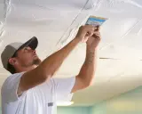 Mann streicht eine Styropordecke mit einer Spachtel.