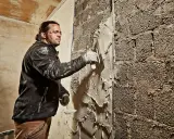 Mann verputzt eine grobe Ziegelwand mit einer Kelle. Der Putz wird gleichmäßig aufgetragen, um die Oberfläche zu glätten.