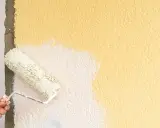 Hand streicht mit einer Farbrolle über eine raue, gelbe Wand. Der Putz wird gerade weiß gestrichen.