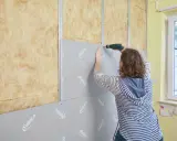 Frau verkleidet Wand mit Rigipsplatten und Schrauber. Isolierung ist sichtbar.