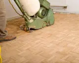 Mann bearbeitet einen Parkettboden mit einer Schleifmaschine. Der Boden schleifen ist ein wichtiger Schritt zur Erneuerung.