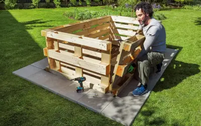 Mann baut ein **Hochbeet aus Paletten** im Garten. Werkzeug und Material liegen bereit.